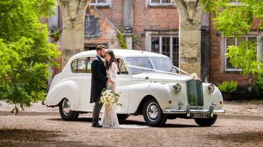 Cream Classis Austin Princess Limousine dressed with Ivory Ribbons and bow across its front bonnet. Bride holding a large bouquet of fresh flowers is standing ion front of the vehicle held by a Groom in a dark great Tuxedo suit.