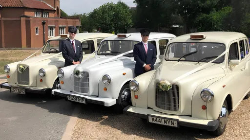 Three Classic Taxicabs decorated with matching ribbons and bows parked next to each others. Two of the Taxis are Ivory coloured and the middle one is in White colour. Two fully uniformed chauffeurs with suit and hat standing in the middle of the vehicles.