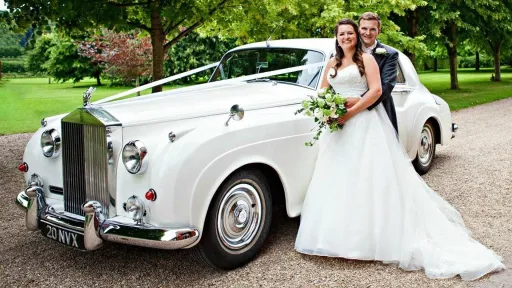 A White classic Rolls-Royce Silver Cloud with single headlights decorated with white ribbons. Bride and Groom standing by the vehicle holding each others.