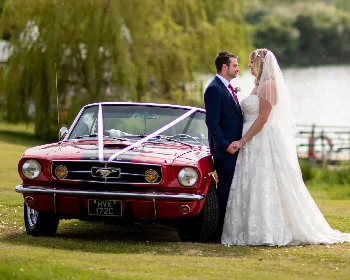 Bride and groom beside a red classic Ford Mustang decorated with wedding ribbons