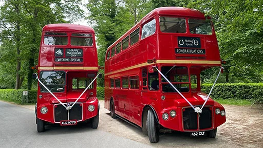 Two vintage red double decker routemaster buses parked side-by-side decorated with white wedding ribbons and bows