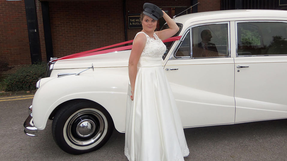 Bride standing n front of the car with hat on. Car is decorated with Red Ribbons