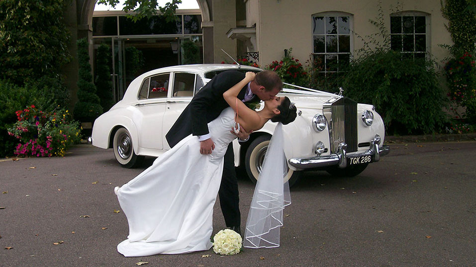 Groom kissing bride in front of their white rclassic rolls-royce