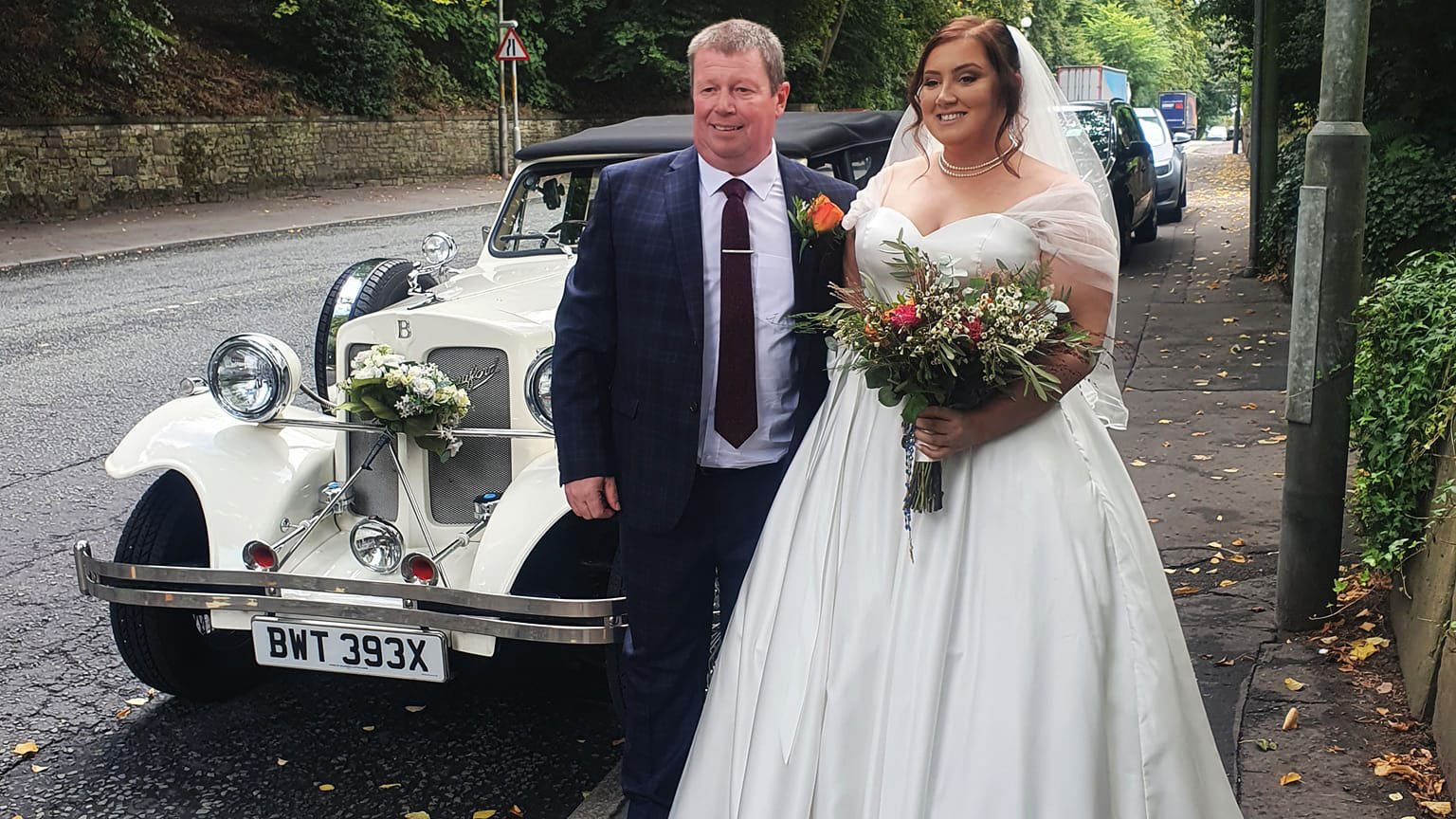Bride with her bridal bouquet and her father in front of the vintage car