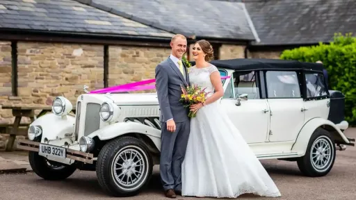White vintage Beauford with whitewall tires, pink ribbon decoration on bonnet and black roof outside wedding venue in Pembrokeshire. Bride and groom standing next to the car with