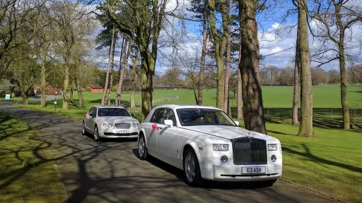 Modern White Rolls-Royce Phantom followed by a White Bentley Flying Spur both dressed with Red bow on the door handling driving through the long entrance at the venue in Pembrokes