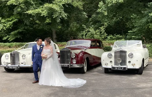 Bride and Groom posing in front of three classic cars they've hired for their Surrey wedding. Each cars are dressed with White Ribbons