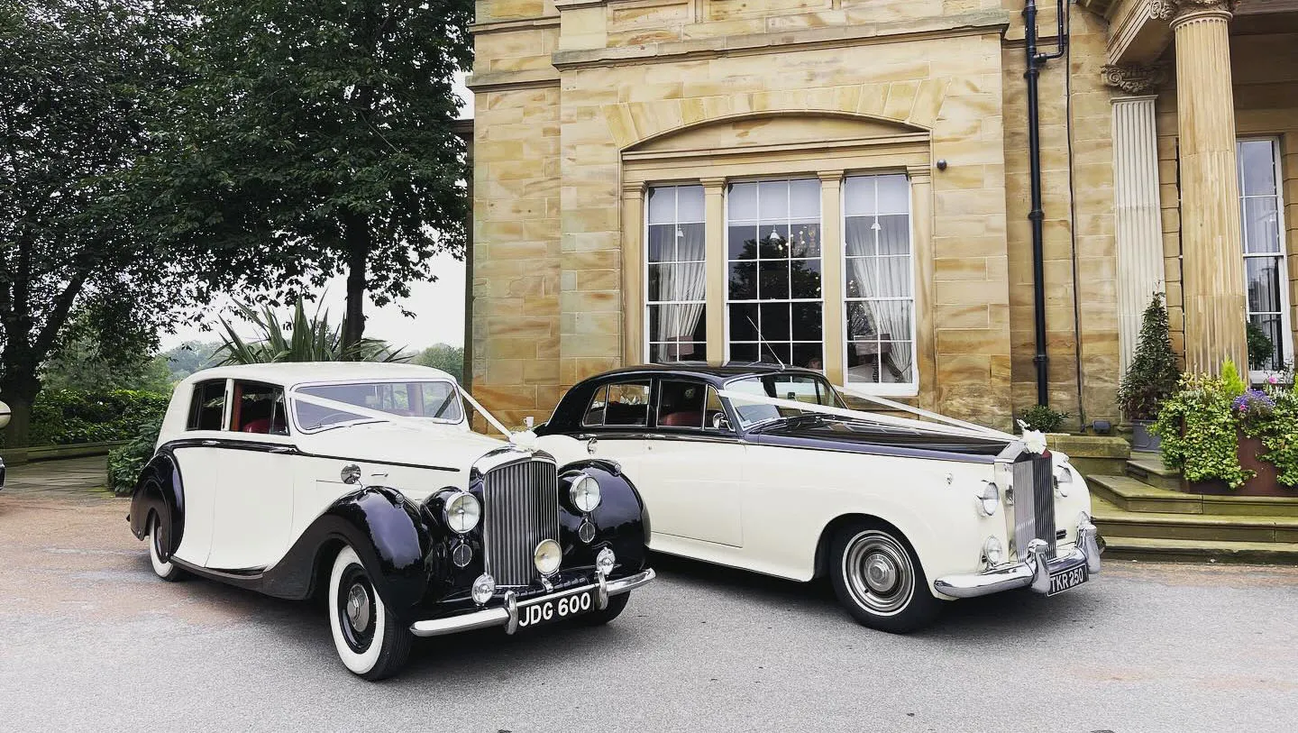 Two classic cars in Black & Ivory with matching White ribbons and bows parked in front of a wedding venue in NewCastle-upon-Tyne.