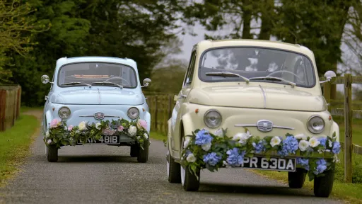 Two Classic Fiat 500 decorated with Wedding flowers on bumper following each other on Lancashire road. Front car on the right is ivory and the rear classic Fiat on the left is in