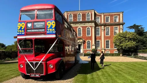Vintage Red Routemaster bus dropping guests off at a Kent wedding venue seen in the background. Bus is decorated with White ribbons and displays vintage advertising on its side.