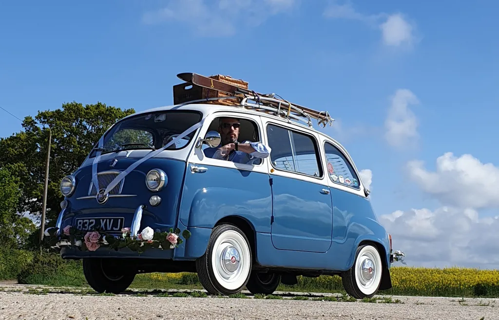 Classic Blue and White Fiat multipla Limousine decorated with flowers on its front bumper and White Ribbons at the front. Chauffeur is inside the vehicle looking out from the side window.