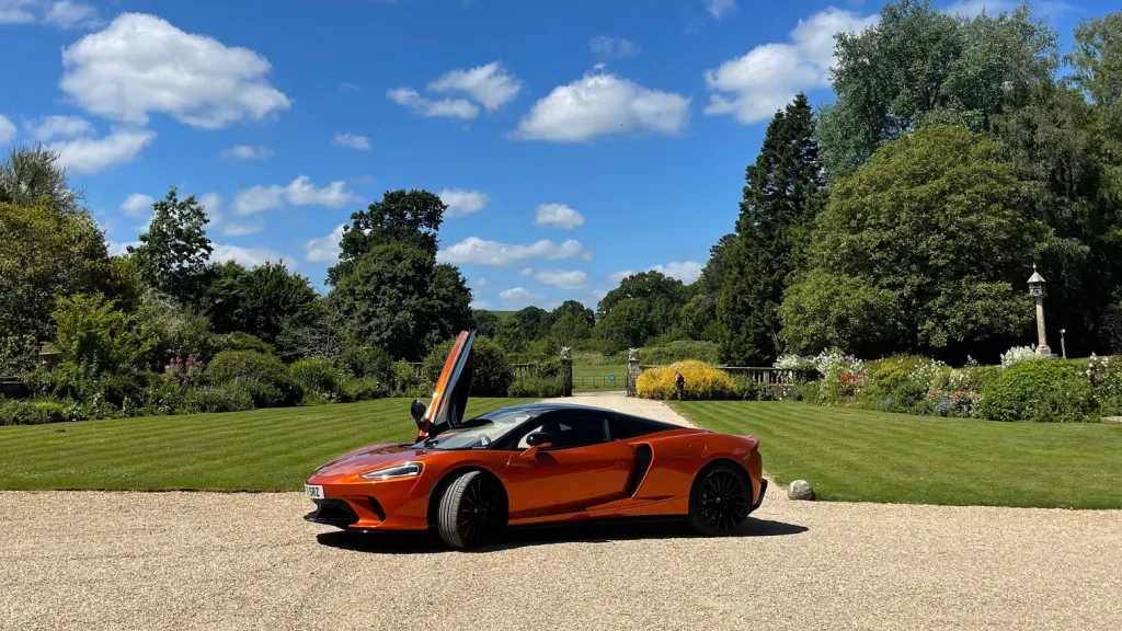 Burnt Orange McLaren GT in front of wedding venue. Gull Wing Door is open on right side