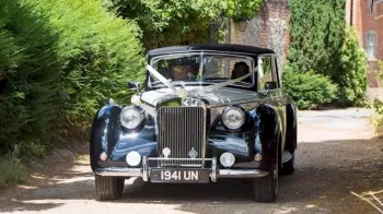 Full front view of Viontage Royale Wedding car decorated with with Ribbons entering wedding venue. Chauffeurs and both passengers can be seen through the windows