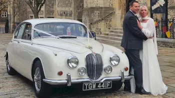 Front view of Mk2 Jaguar with pale pink Ribbons accross the front of the vehicle. Both Bride and Groom are standing next to the car posing for their photographer