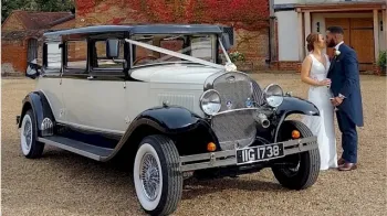 Black & White vitage Bramwith dressed with white ribbons, white wall tires with bride and groom standing on the left of the vehicle kissing