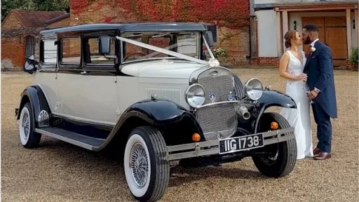 Black & White vitage Bramwith dressed with white ribbons, white wall tires with bride and groom standing on the left of the vehicle kissing
