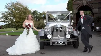Full front view of imperial car with white ribbons and Bride and groom standing on either side of the vehicle. Bride is wearing a large white dress and holding a bouquet of flowers