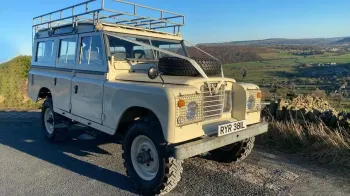 Front Side View Cream Classic Landrover with roof rack, wheel on bonnet and white ribbon decoration accross the front