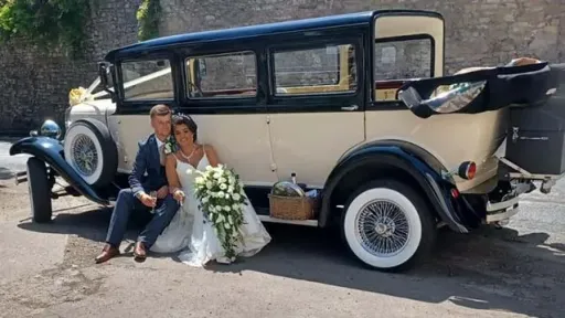 Bride and Groom sitting on the Wheel Arche of a Vintage Bramwith posing for photos. Bride is holding a big Bouquet of flowers