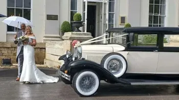 Bride and Groom having their photos taken in front of a Vintage BRamwith Limousine. Groom is holding a white umbrella and Bride is holding flowers