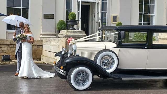 Bride and Groom having their photos taken in front of a Vintage BRamwith Limousine. Groom is holding a white umbrella and Bride is holding flowers