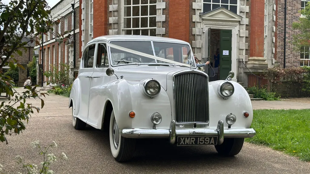 Front view of Classic Austin Princess decorated with white ribbons across its bonnet leaving a wedding venue in Essex