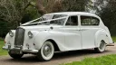 Front Left side view of classic Austin Princess Limousine decorated with white ribbons in a park with green tree in the background