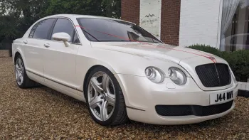 Front Right Side view of White Bentley Flying Spur showing the Chrome Alloy Wheels and double twin headlights in front of a wedding venue in Bedford