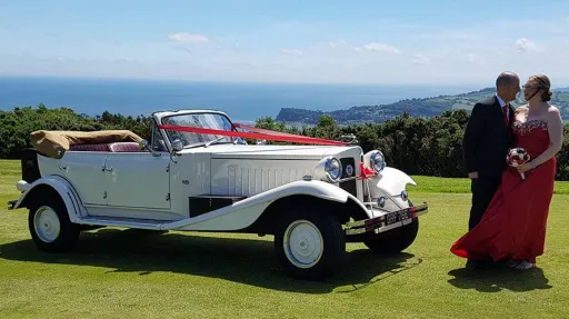 Beauford 4 Door Convertible on top of cliff decorated with burgundy ribbons with bride and groom standing in front of the vehicle