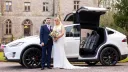 Bride and Groom stansing in front of a white Tesla X with Gull-wings open in front of a wedding venue