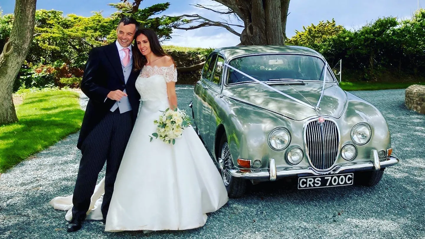 Classic Jaguar MK2 with Bride and Groom standing in front of the vehicle smiling. The Bride is holding her bridal bouquet in her left hand. Vehicle is parked in the garden of a lo