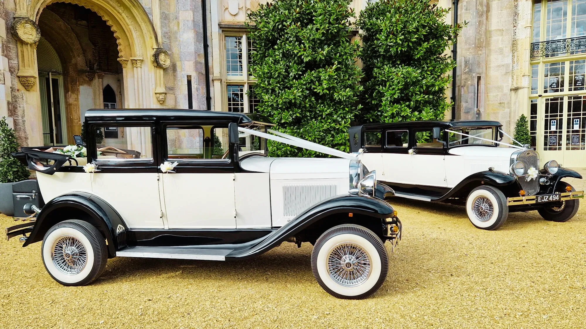Two matching vintage wedding cars decorated with matching white ribbons. Vehicles are parked at the front entrance of a local wedding venue in Sherborne.
