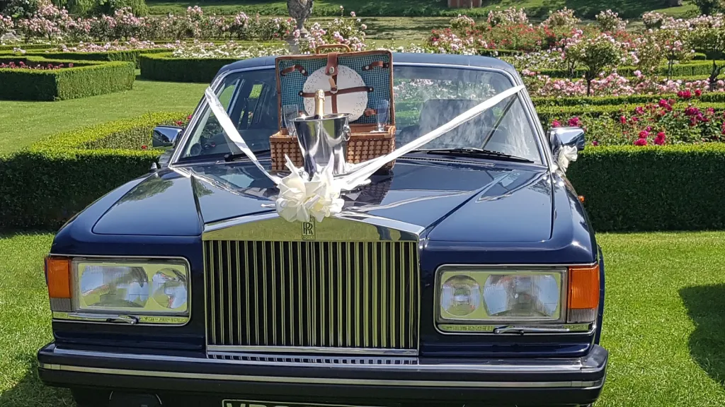 Full front view of blue rolls-royce with white ribbons and bow and a picnic basket with champagne glasses on top