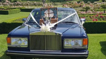 Full front view of blue rolls-royce with white ribbons and bow and a picnic basket with champagne glasses on top