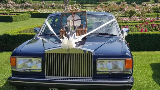 Full front view of blue rolls-royce with white ribbons and bow and a picnic basket with champagne glasses on top