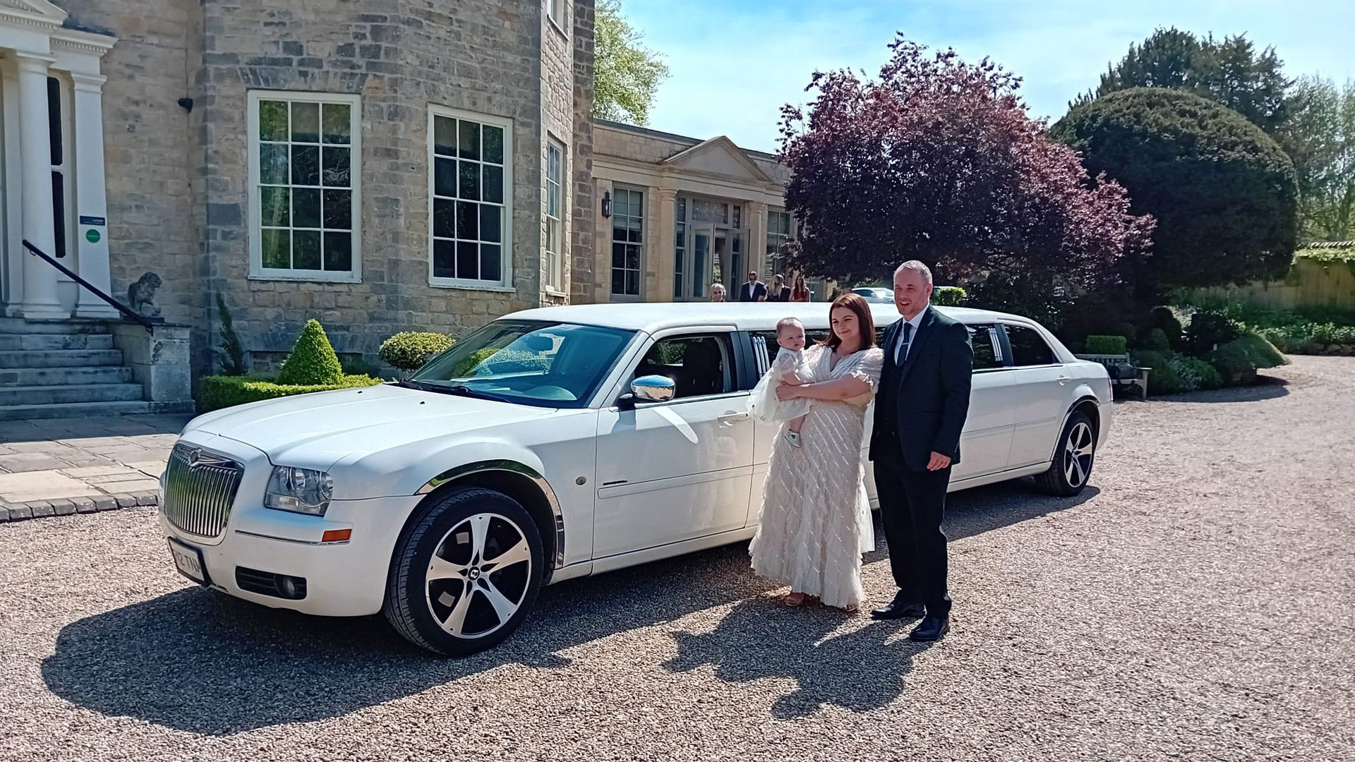 Chrysler 300c White Limo in front of a wedding venue with Bride and Groom standing by the vehicle. The Bride is holding her little infant baby in her arm.