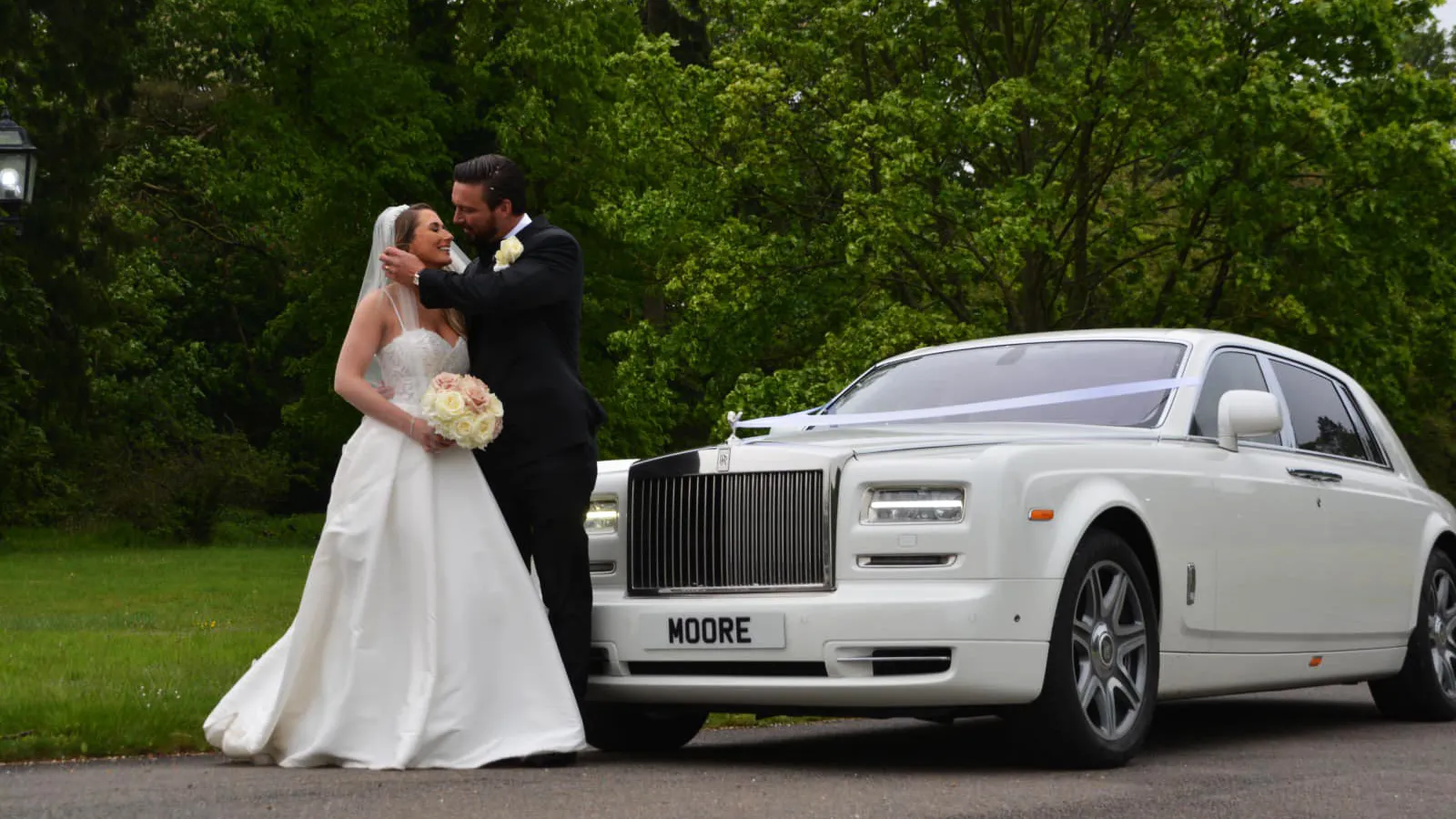 White Rolls-Royce Phantom decorated with white ribbons with Bride and Groom standing in front of the vehicle
