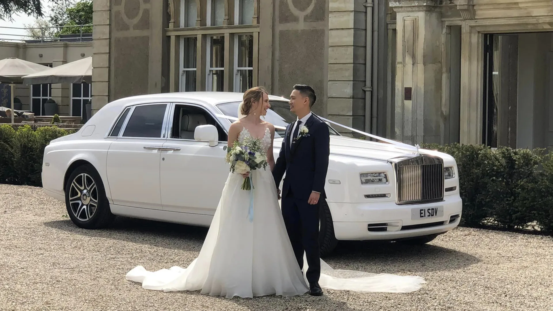 White Rolls-Royce Phantom decorated with white ribbons with Bride and Groom standing in front of the vehicle.