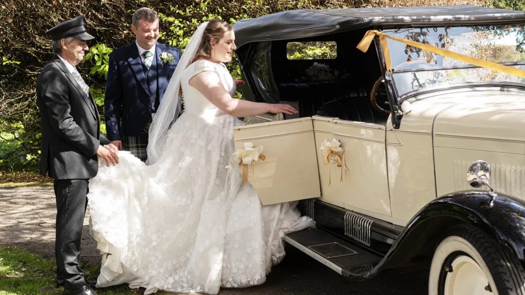Chauffeur holding the bride's dress and helping her to get into the vintage car