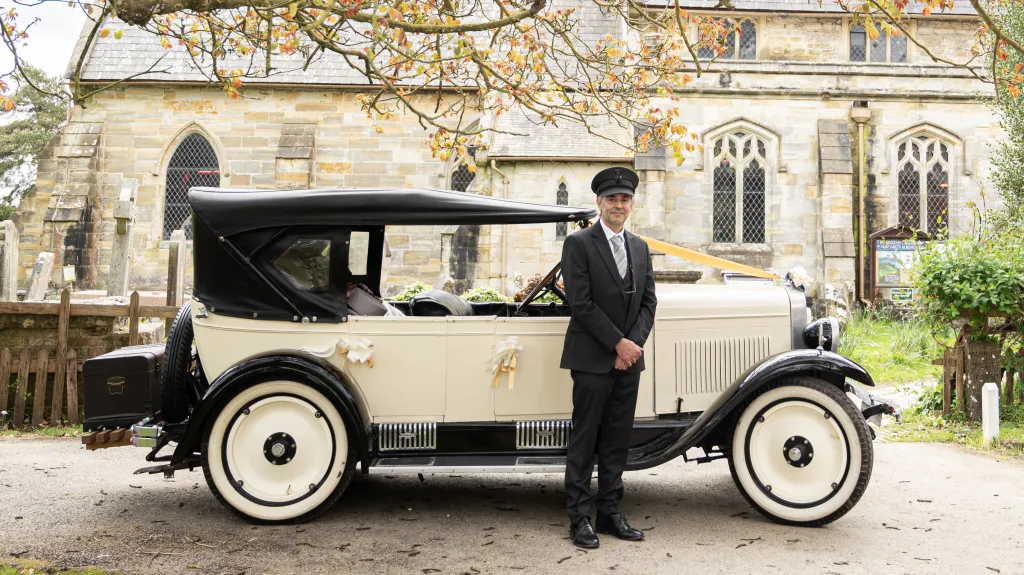 Fully Uniformed chauffeur standing in front of an American chevrolet. Church in the background