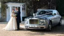 Bride and Groom holding each others in front of their wedding venue with Modern Rolls-Royce Phantom in silver dressed with ribbons on the side