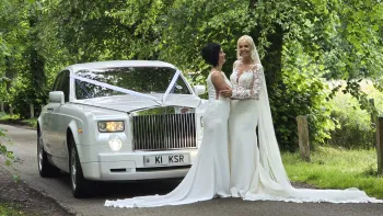 LGBT Gay Wedding with two brides in their wedding dress holding each others and smiling in front of a white rolls-royce phantom decorated with white ribbons