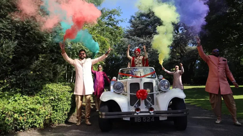 Asian wedding with groom standing inside a vintage beauford with roof down and groom's men walking next to the vehicle wetting off colourful smoke flares