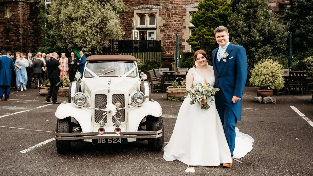Bride and Groom standing next to a Vintage Beauford convertible. Wedding guests can be seen in the background entering the Reception venue.