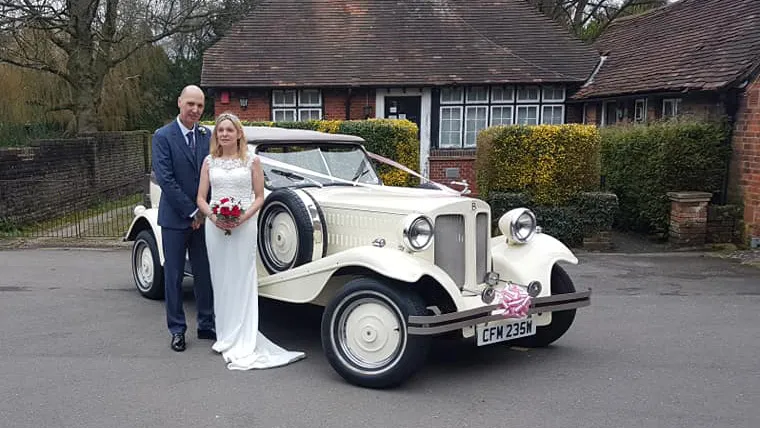 Bride and Groom standing in front of a Vintage Beauford Convertible dressed with light pink ribbons and large bow on front bumper. Bride is holding a bouquet of flowers.