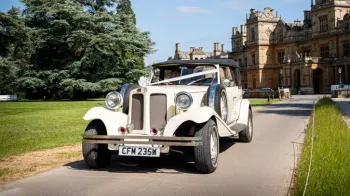Vintage Beauford Convertible with spare wheel mounted on side of the vehicle decorated with white ribbons in front of a wedding venue in Cotswolds.
