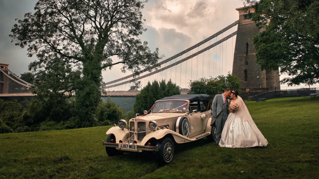 Ivory vintage Beauford parked in front of Clifton Suspension bridge with Bride and Groom kissing next to the vehicle