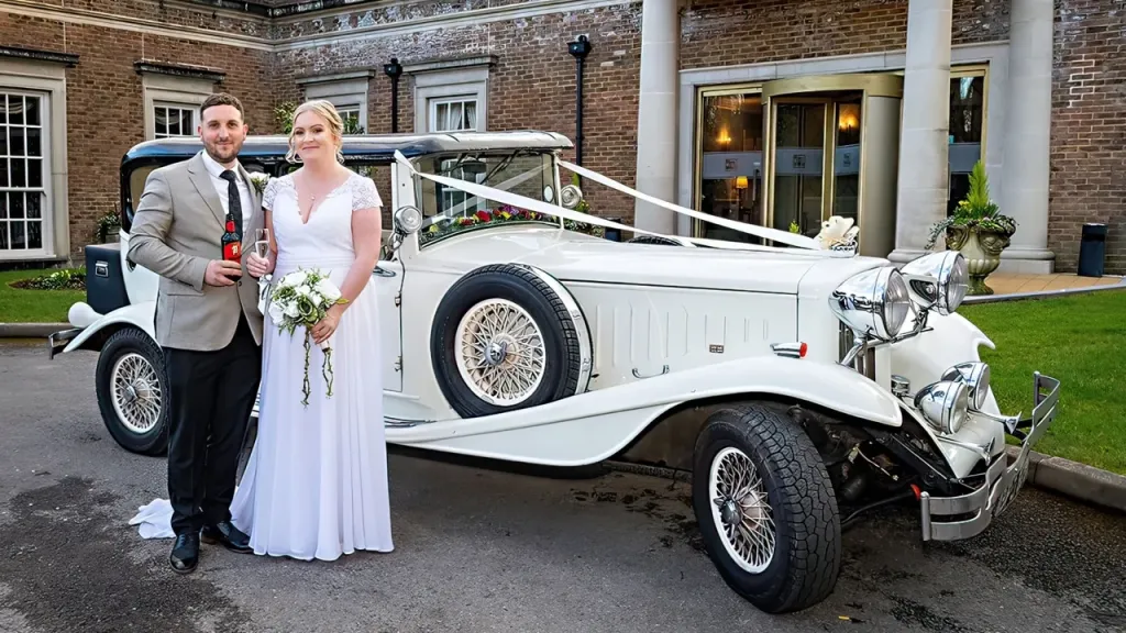 Bride and Groom standing in front of a vintage Beauford decorated with white ribbons outside their wedding venue.