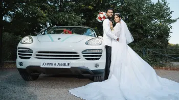 Bride and Groom standing next to White porsche Cayenne decorated with light blue ribbons. Bride is wearing a large white dress and groom a suit with black trousers and white jacket.