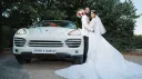 Bride and Groom standing next to White porsche Cayenne decorated with light blue ribbons. Bride is wearing a large white dress and groom a suit with black trousers and white jacket.